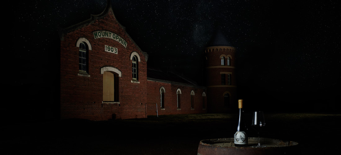 Wine bottles and glass on top of barrel with the winery facade as background at night.